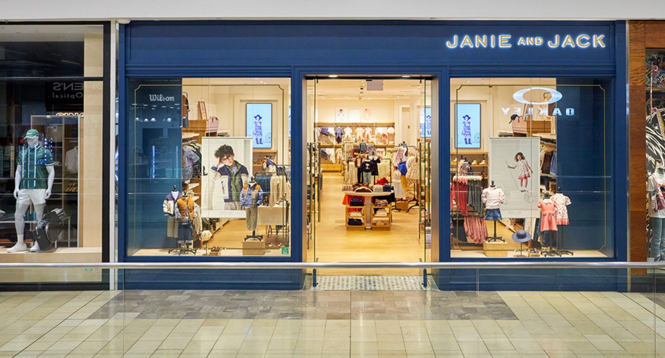 Wide shot of a Janie and Jack mall storefront from across the concourse, multiple digital window displays visible.