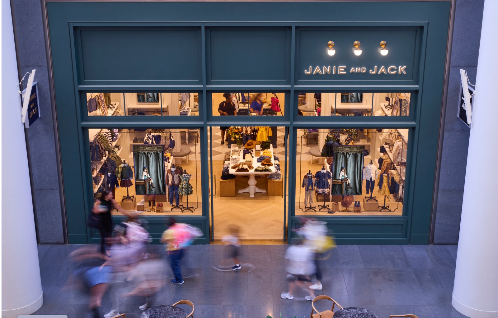 Wide shot of a Janie and Jack mall storefront with synchronized digital window displays and shoppers walking past