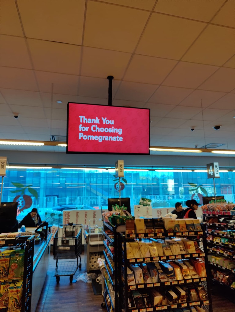 Grocery store aisle with a hanging digital screen reading “Thank You for Choosing Pomegranate” above shoppers and shelves.