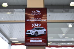 Vertical digital sign in a car showroom showing a Toyota of Olympia Highlander lease offer.