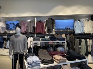 Mannequin and folded menswear tables with two in-store digital screens above clothing racks.