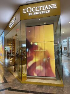 L’Occitane corner storefront with a tall multi-panel video wall playing a brand advertisement.