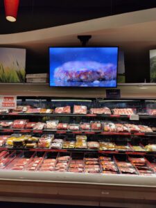 Grocery meat department with a widescreen display above refrigerated meat cases.