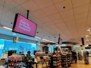 Grocery store interior with a hanging digital screen above aisles and checkout displays.