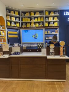 L’Occitane checkout counter with shelves of gift boxes and a large horizontal digital screen behind the cashier.