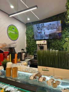 Juice bar counter with a plant wall and a wall-mounted TV showing a production scene above the register area.