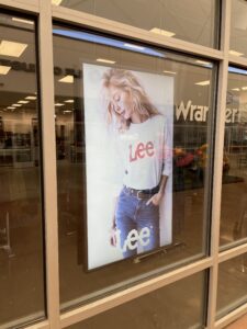 Close view of a vertical digital sign showing a Lee model through a storefront window.